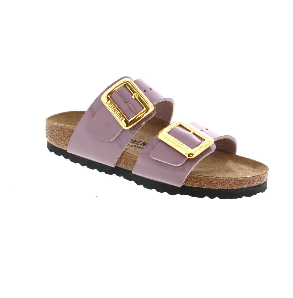 Introducing the Birkenstock Sydney Luxe sandal in Faded Purple. Featuring an oversized statement buckle and shimmery Birko-Flor® material, this sandal adds a touch of shine to any outfit. The contoured footbed provides excellent support for all-day comfort, making it perfect for chasing the sun.